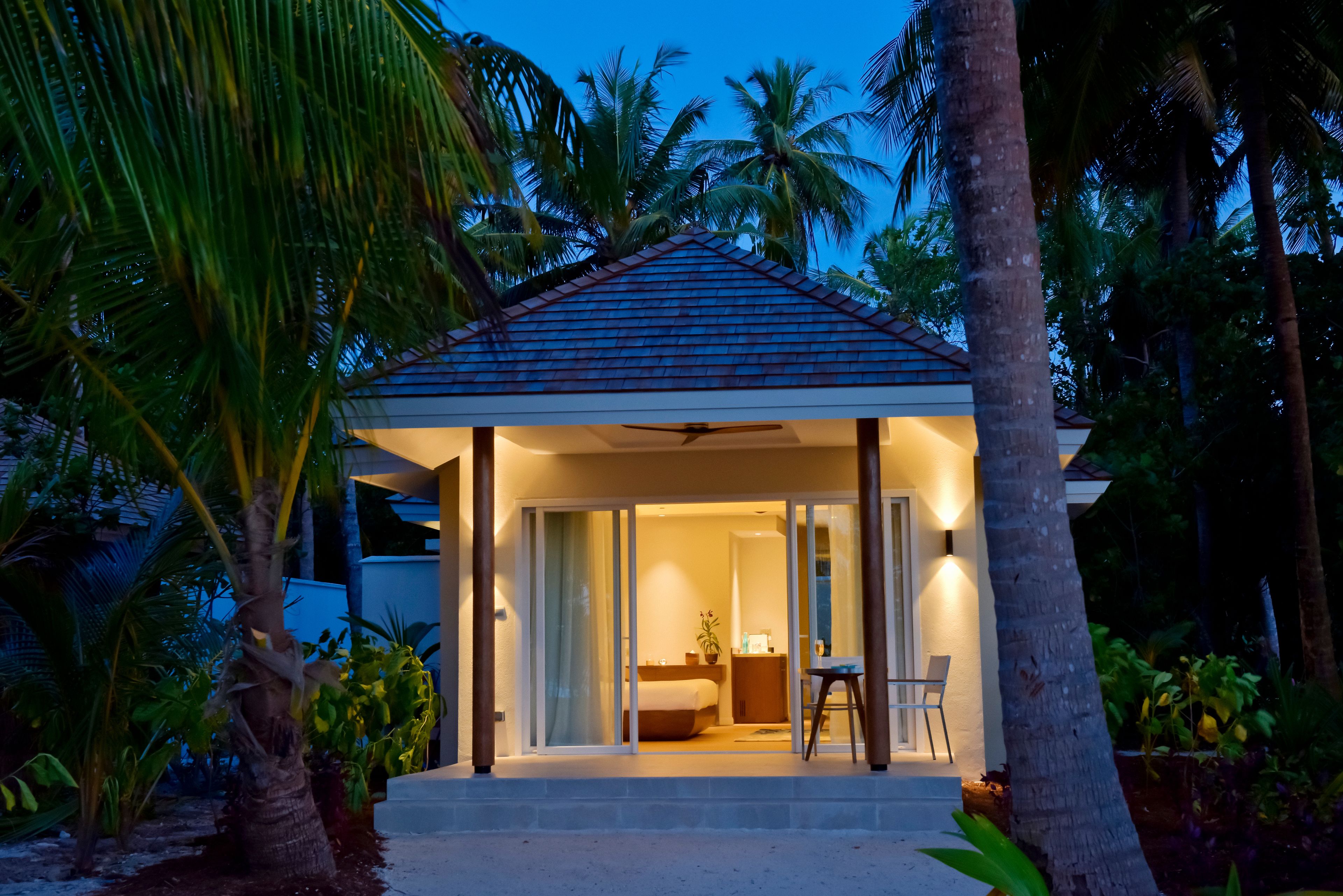 Une maison de style bungalow moderne, dotée d’un porche couvert, d’une chambre avec un lit et une table de chevet, ainsi que d’un coin repas comprenant une chaise et une table.