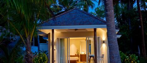 Une maison de style bungalow moderne, dotée d’un porche couvert, d’une chambre avec un lit et une table de chevet, ainsi que d’un coin repas comprenant une chaise et une table.
