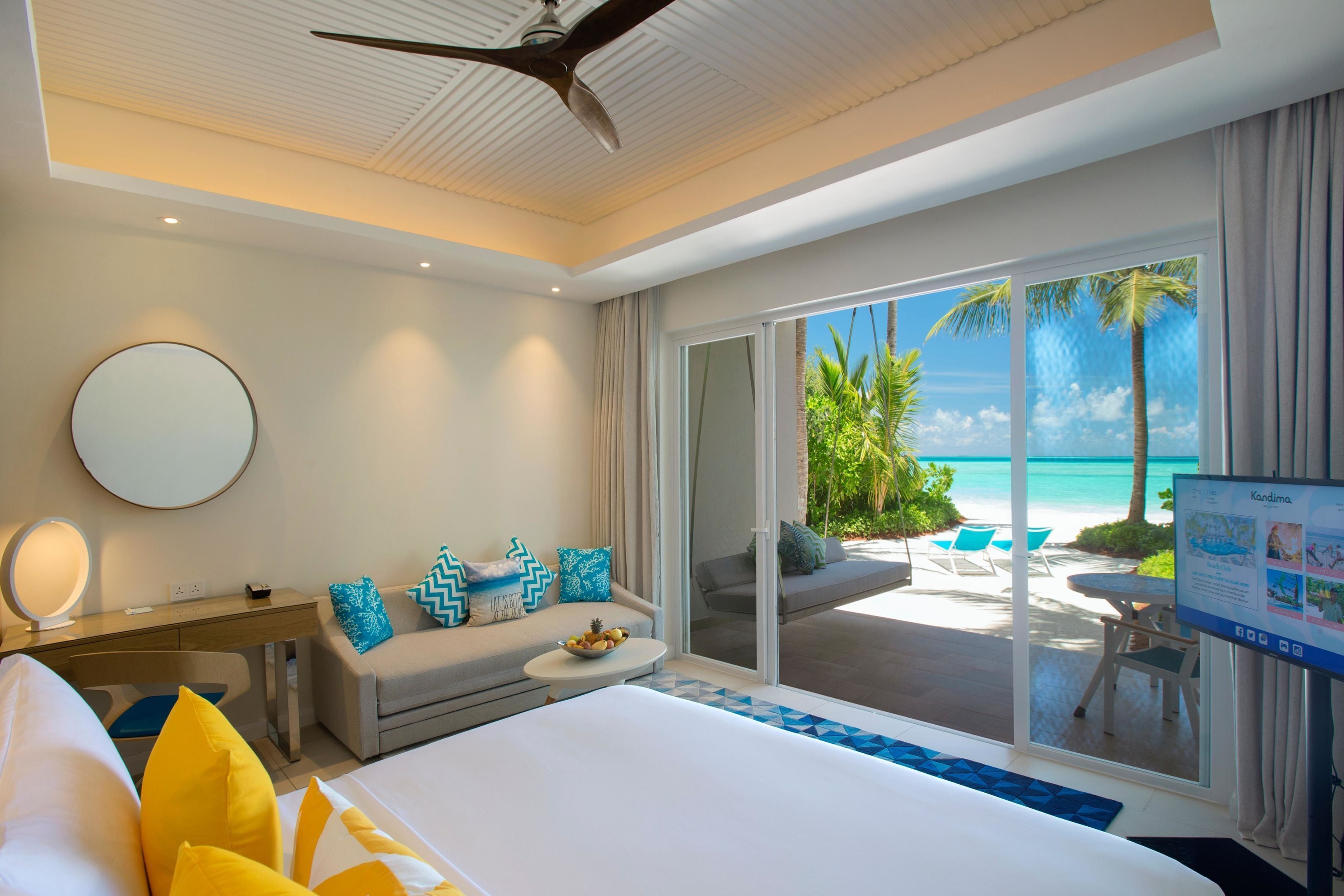 A hotel room with a bed, a desk with a mirror, a sofa with cushions, a small table with fruit, and a view of the beach and palm trees through large sliding doors.