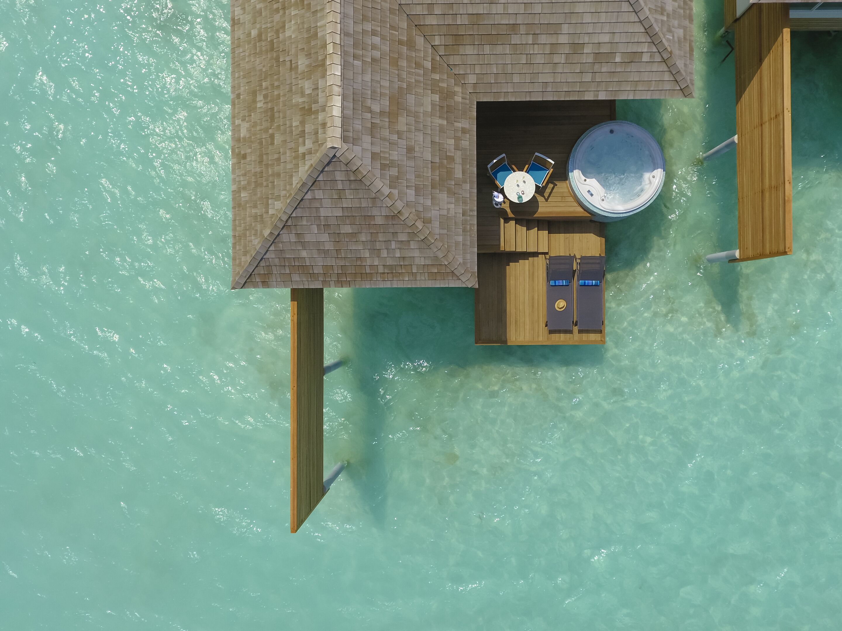 An aerial view of a wooden overwater bungalow with a hot tub, two lounge chairs, and a small table.