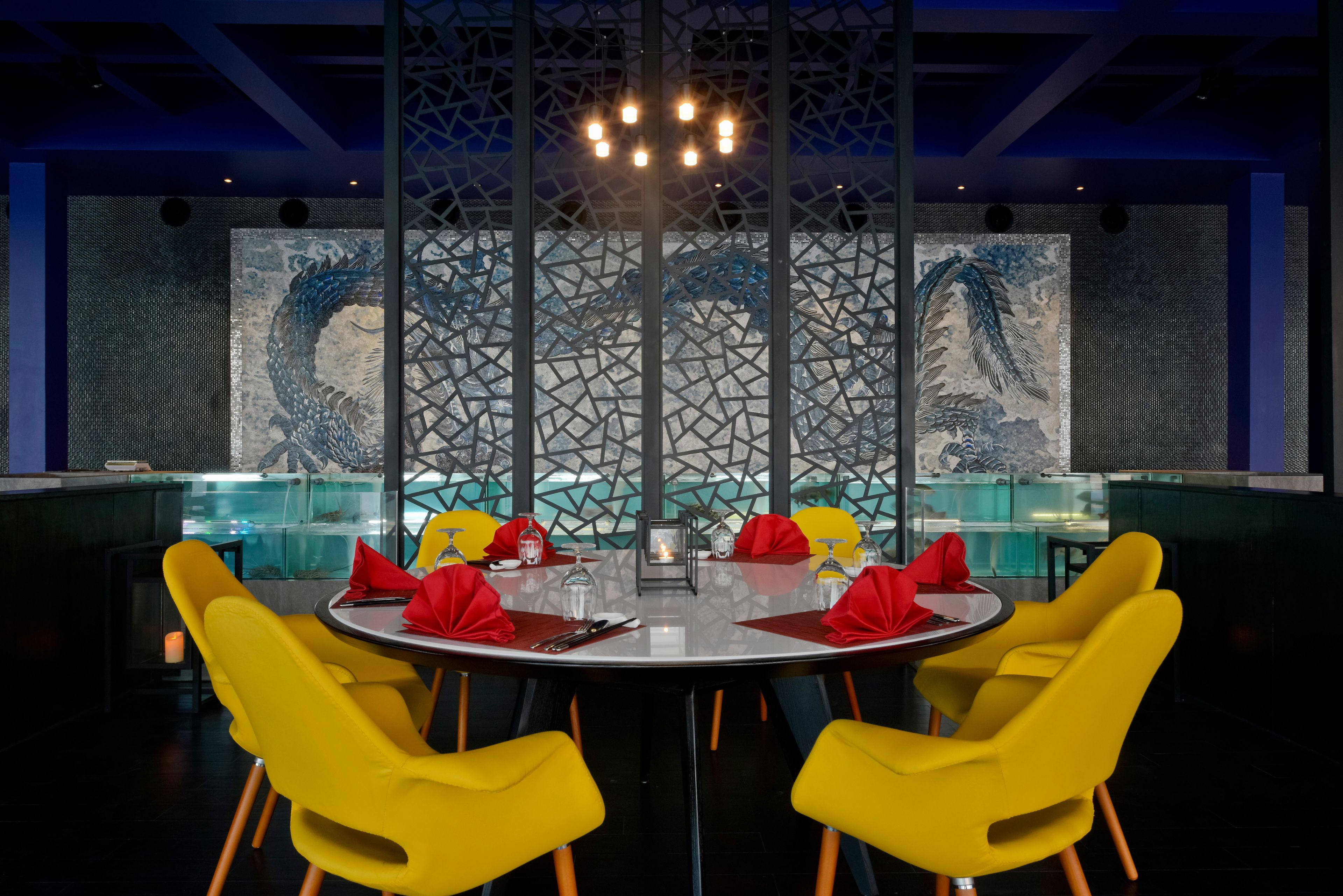 Un comedor con una mesa redonda dispuesta para cuatro personas, sillas amarillas y una pared decorativa con un diseño de dragón.