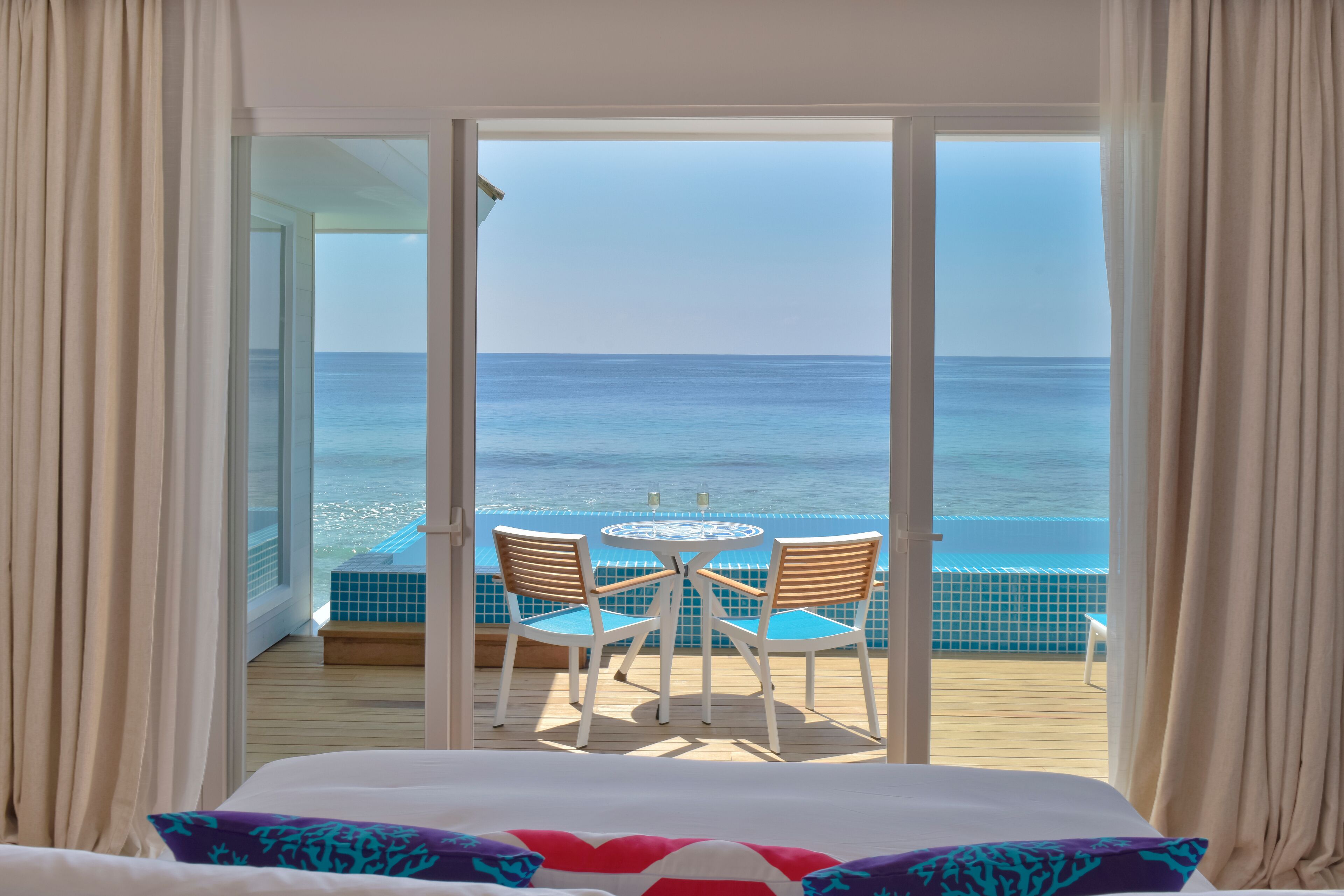 Una habitación con cama, vista al océano a través de amplios ventanales y un pequeño balcón con mesa y sillas.