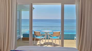 Una habitación con cama, vista al océano a través de amplios ventanales y un pequeño balcón con mesa y sillas.