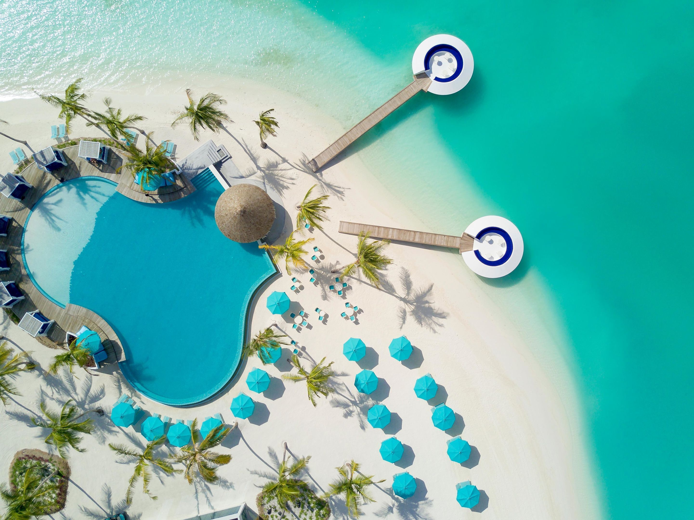 An aerial view of a resort with a central swimming pool, multiple lounge areas with umbrellas, and a beach area with sun loungers and palm trees.