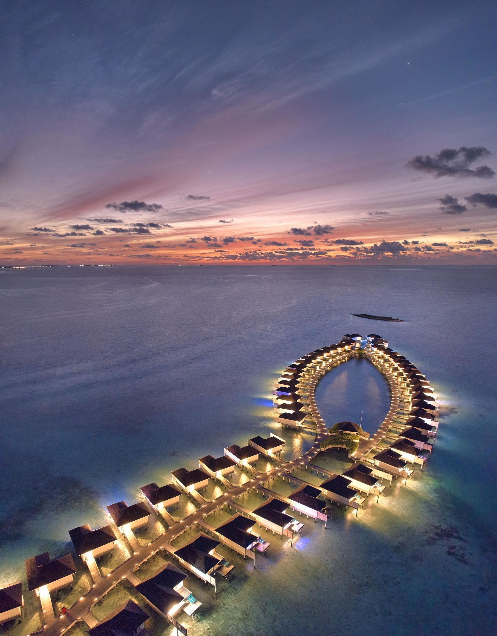 An aerial view of a resort with overwater bungalows arranged in a circular pattern, connected by walkways.