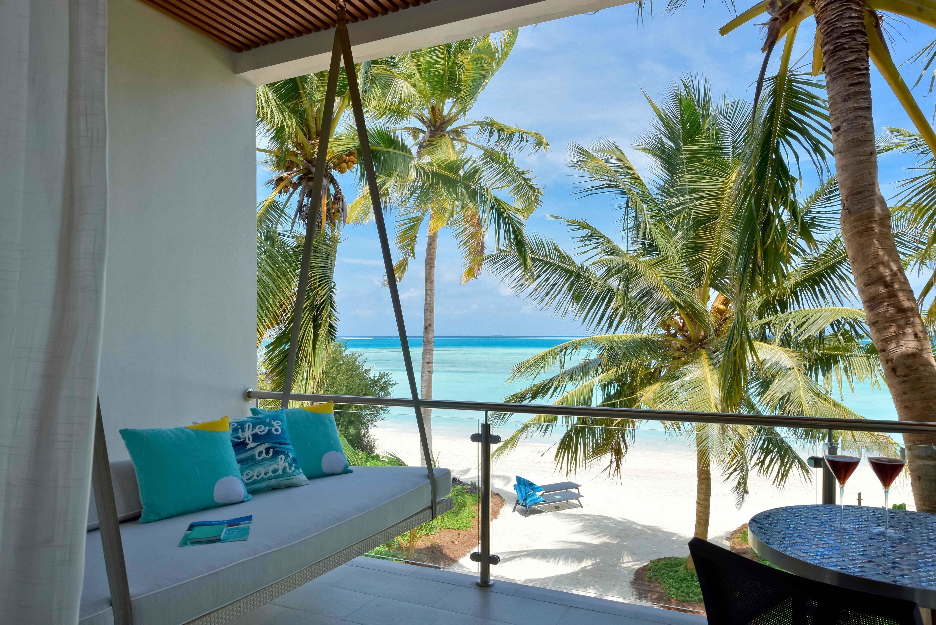 Un balcón con vistas a la playa, palmeras y un área de descanso con sofá y sillones.