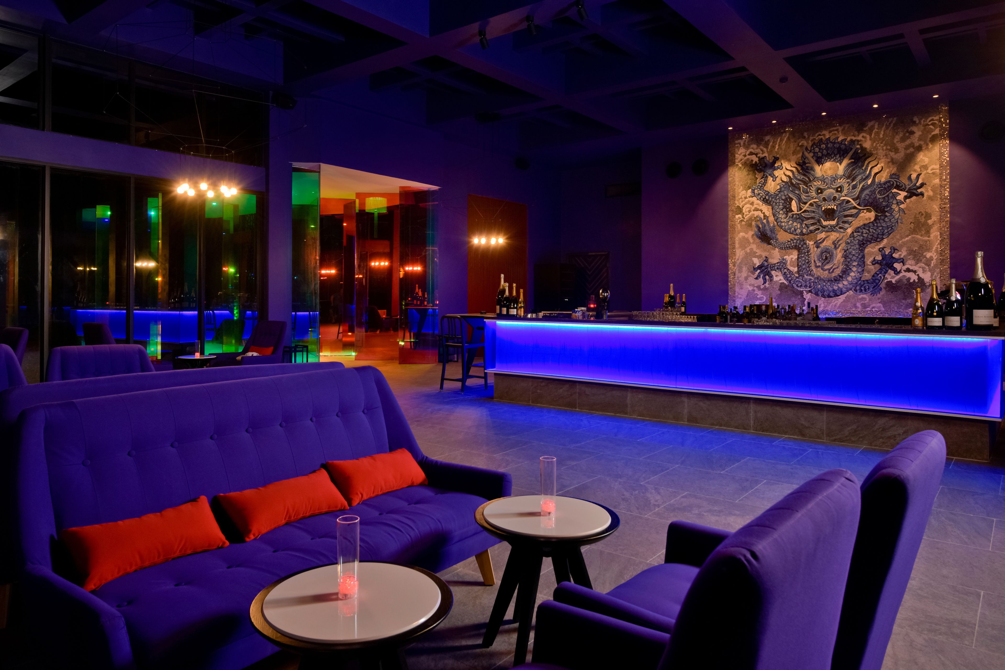 Un espace bar moderne avec un comptoir illuminé de lumière bleue, des sièges violets et une fresque murale décorative.