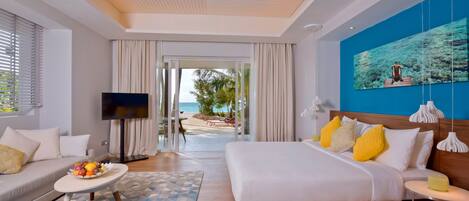 A modern hotel room with a bed, a TV, a dining area, and a view of the beach.
