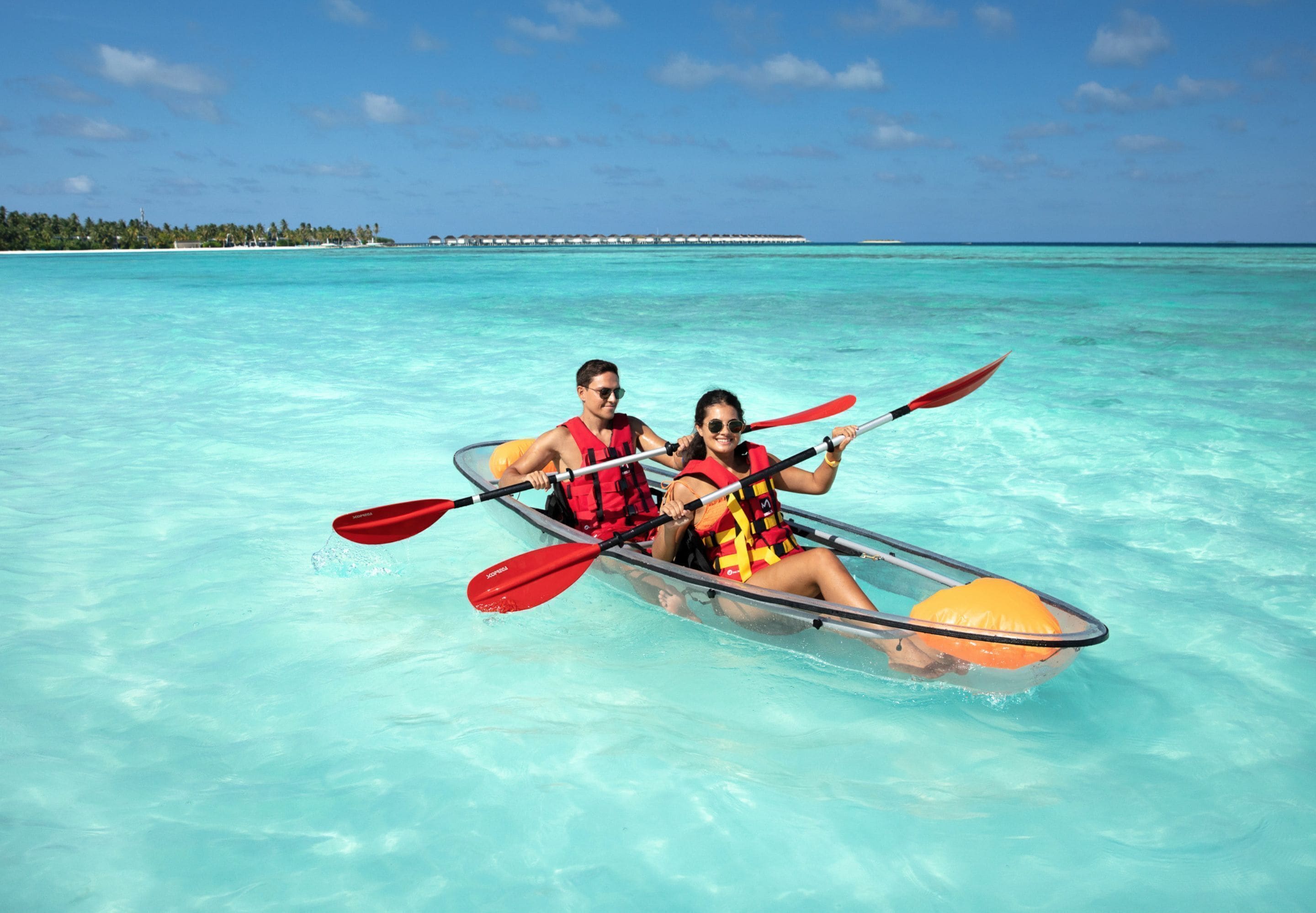 A couple in a transparent kayak on clear turquoise water with a distant island and overwater bungalows.