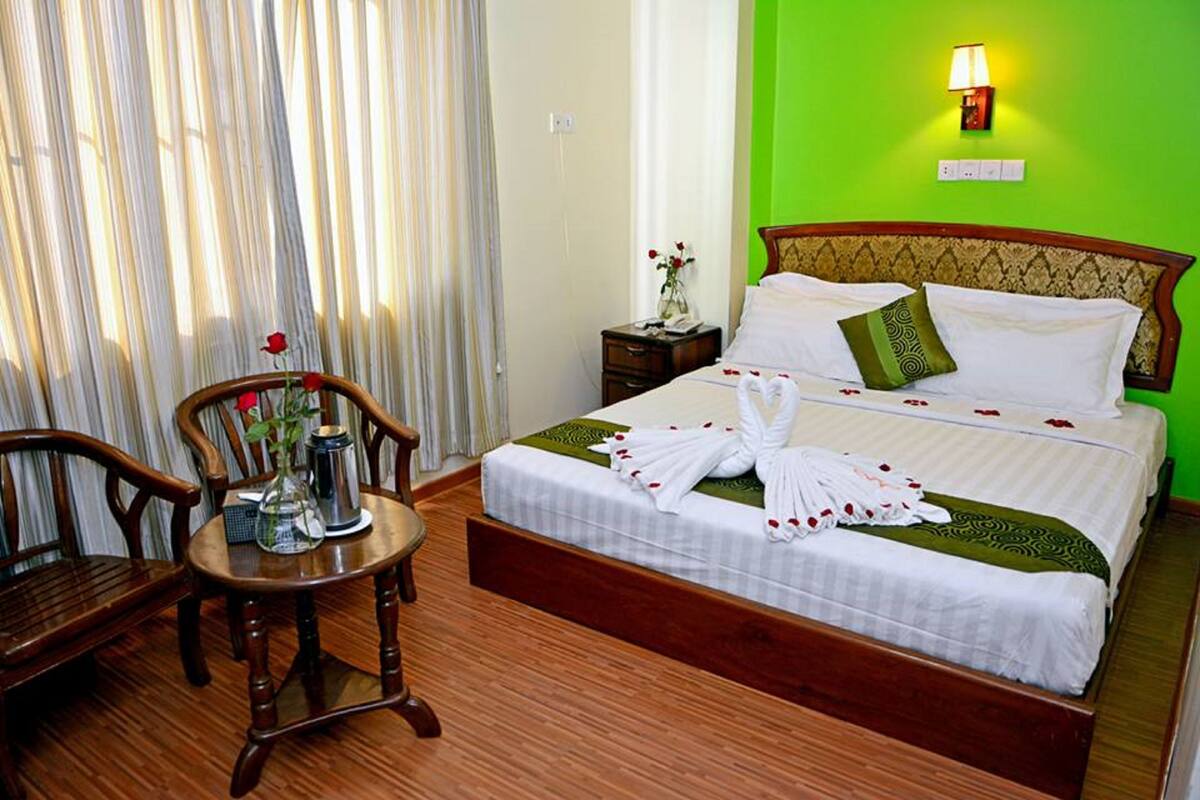 A hotel room with a bed, a wooden chair, a small table with a vase of flowers, and a nightstand with a lamp.