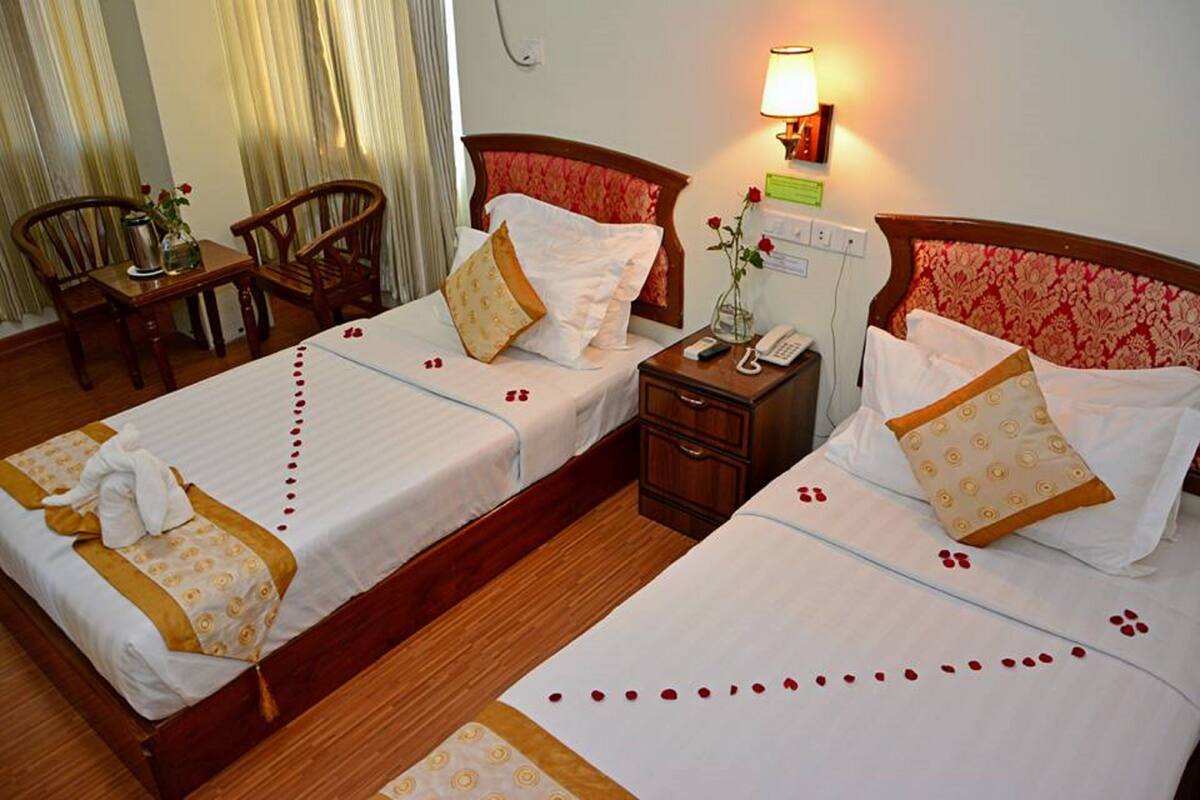 A hotel room with two beds, a bedside table with a lamp, a telephone, and a small table with chairs.