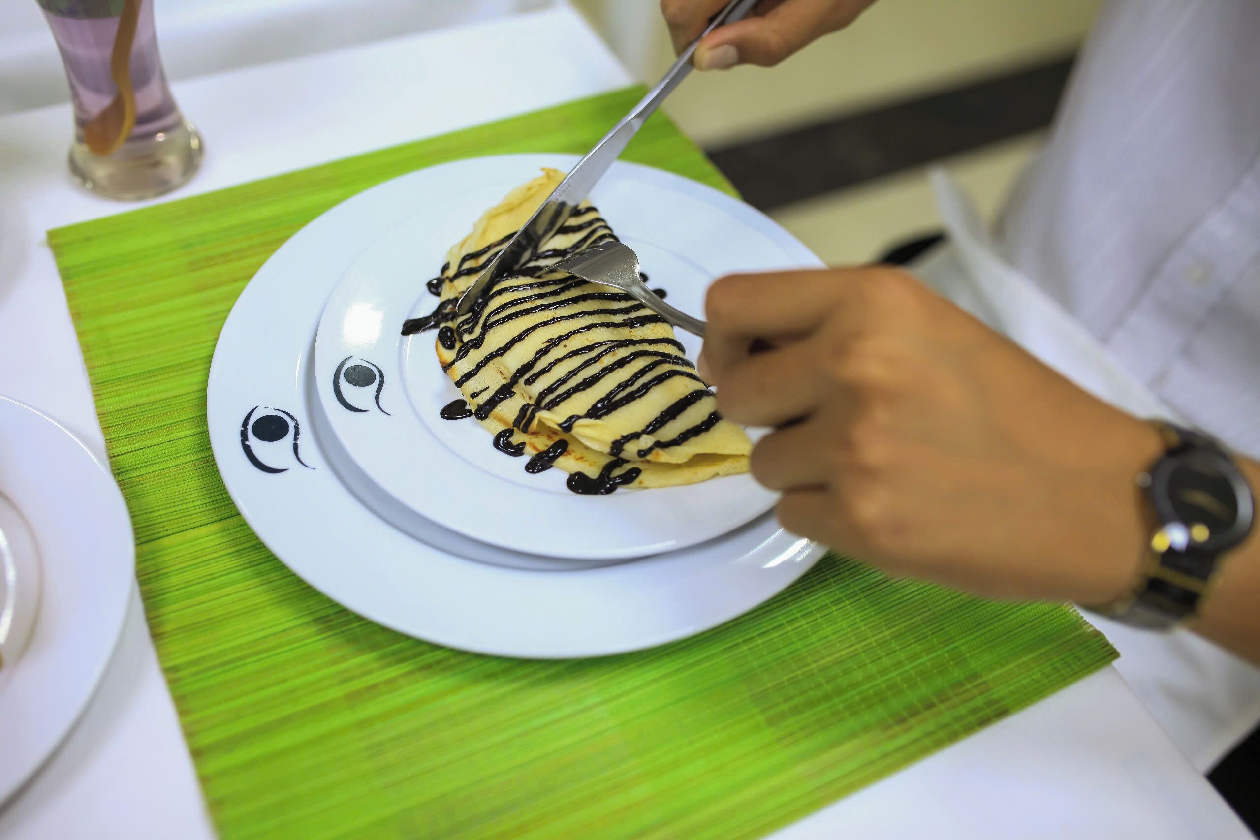 A person cutting a crepe on a white plate with chocolate sauce, placed on a green placemat.