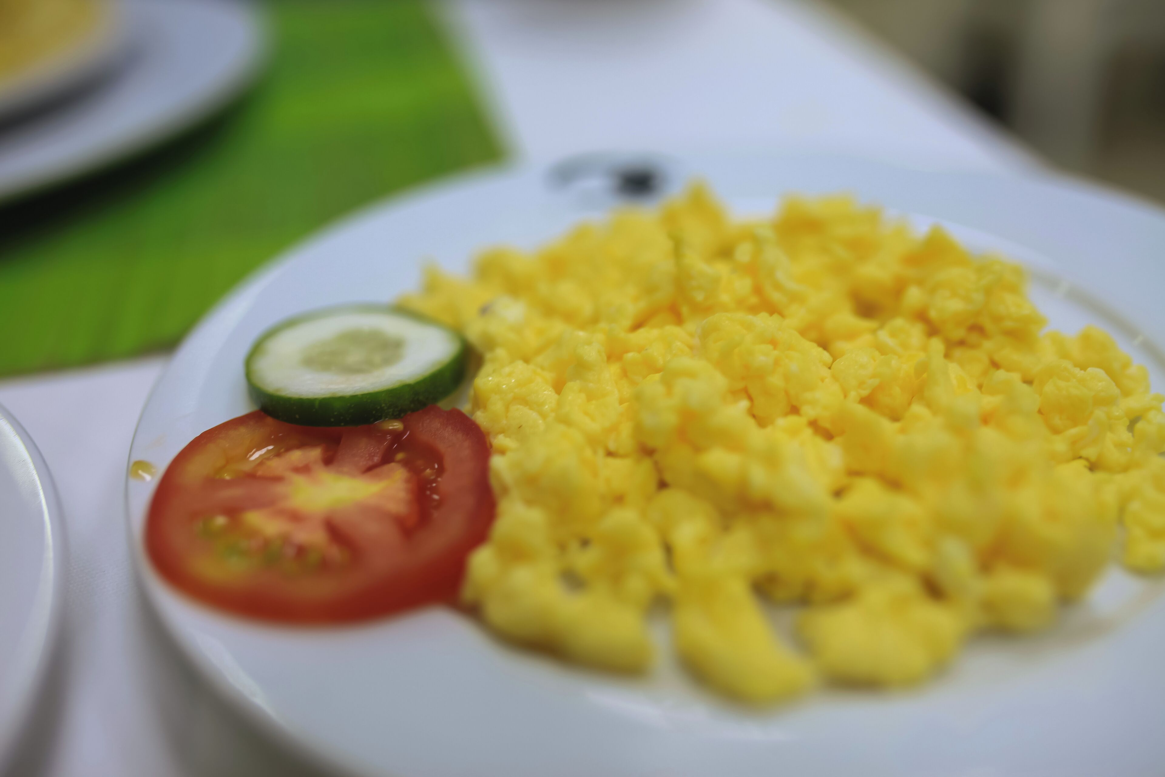 A plate of scrambled eggs with a slice of tomato and a slice of cucumber.