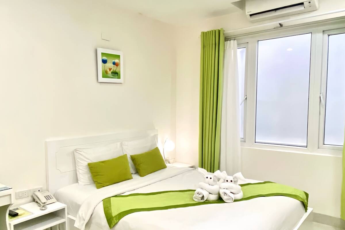 A hotel room with a neatly made bed, a bedside telephone, a small table with a lamp, and a window with curtains.