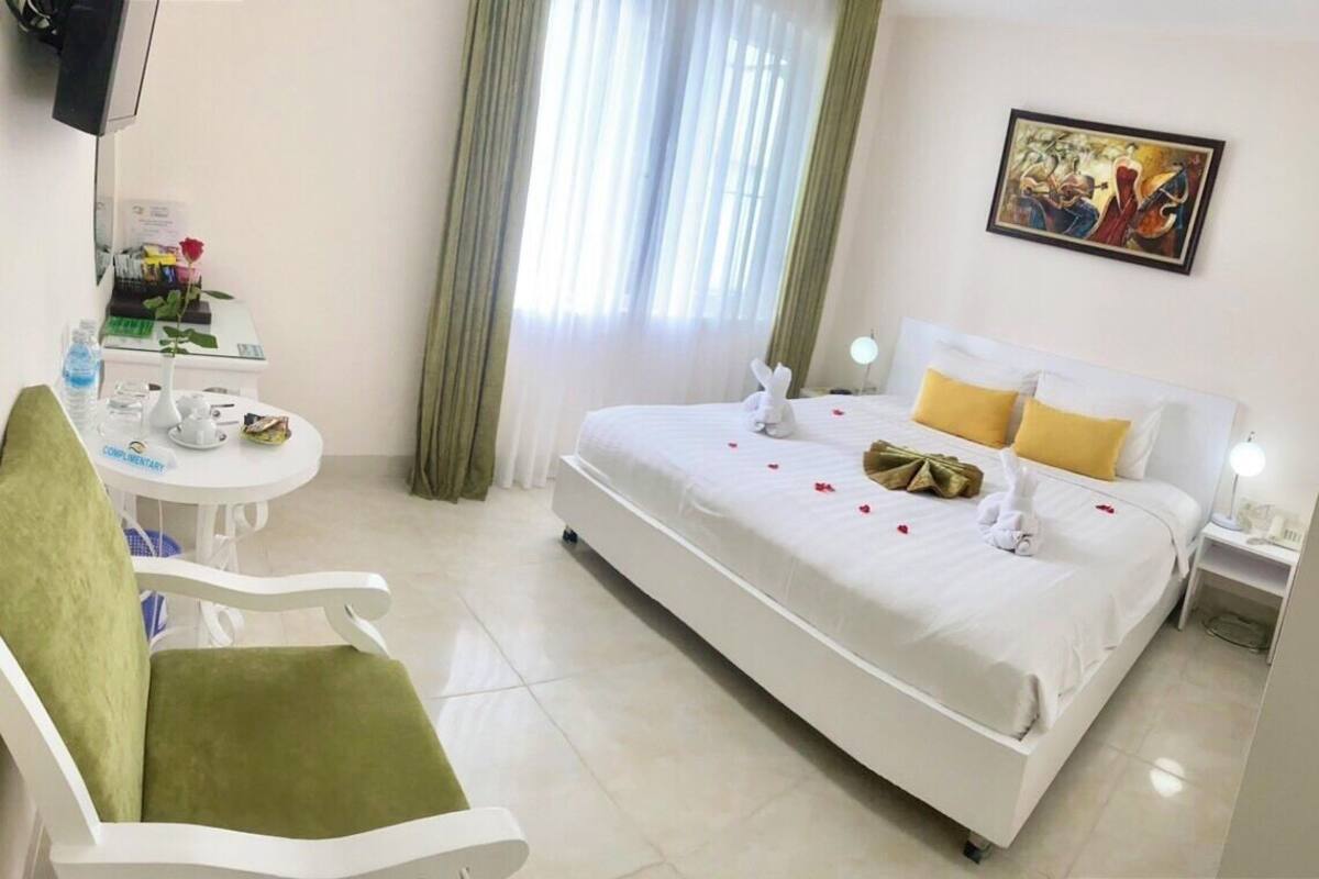 A hotel room with a bed, a chair, a small table with a rose, and a painting on the wall.