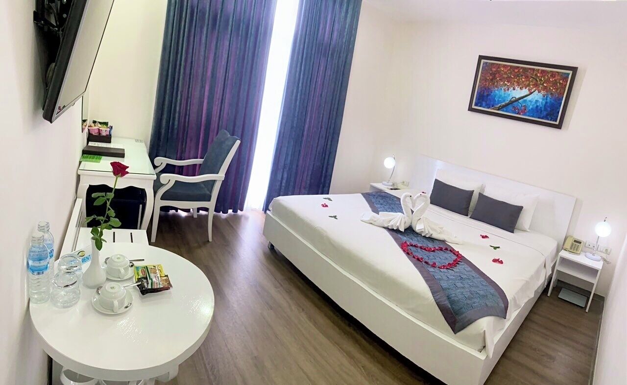 A hotel room with a bed, a desk, a chair, a round table with a vase of flowers, and a painting on the wall.