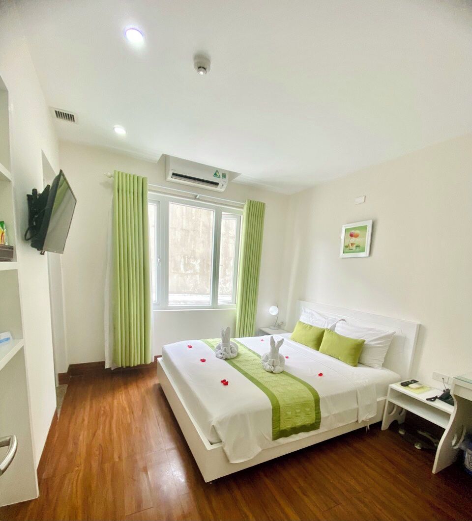 A hotel room with a bed, a TV mounted on the wall, a window with curtains, an air conditioning unit, and a bedside table with a lamp.