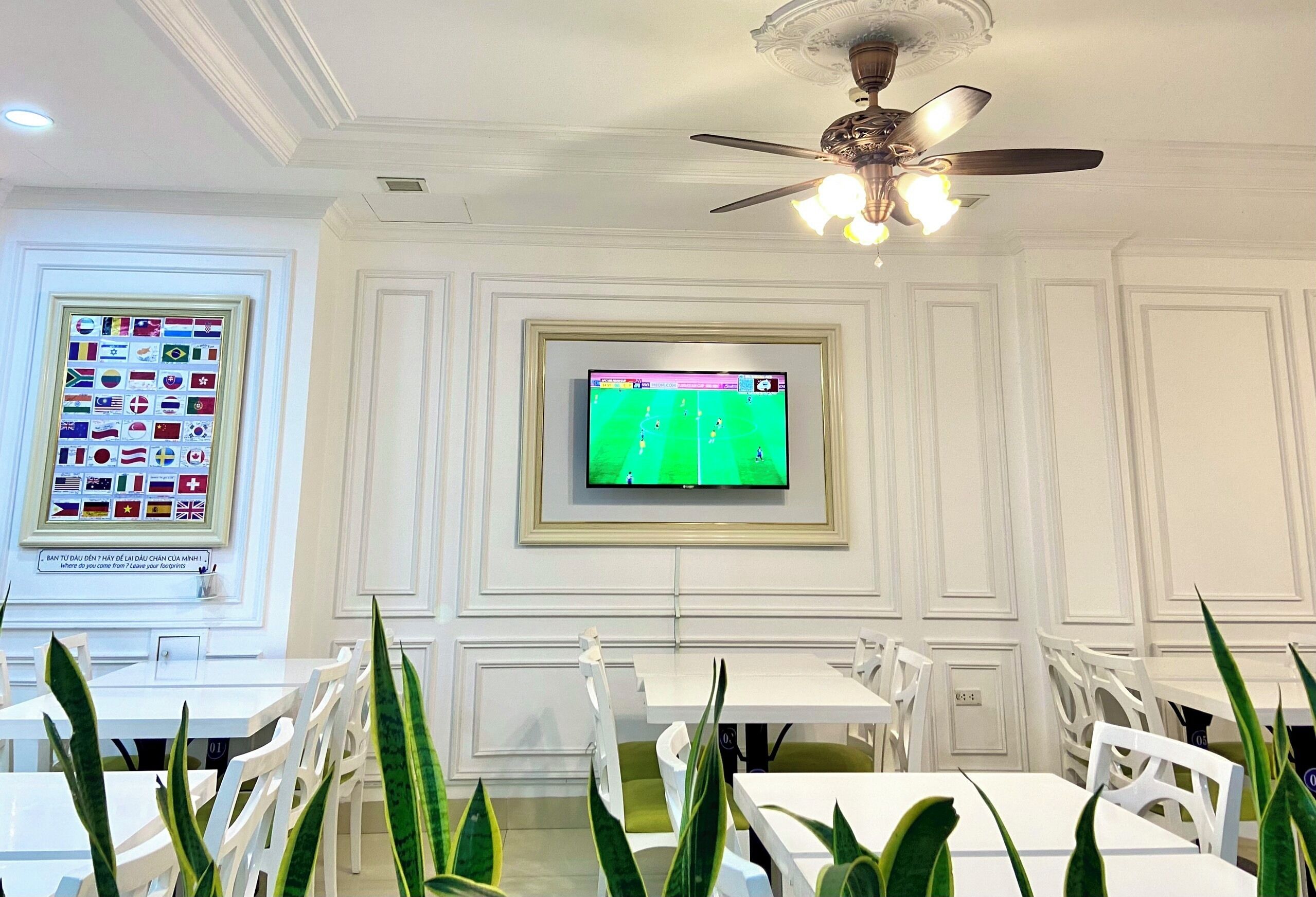 A dining area with white tables and chairs, a ceiling fan, and a television displaying a soccer game.