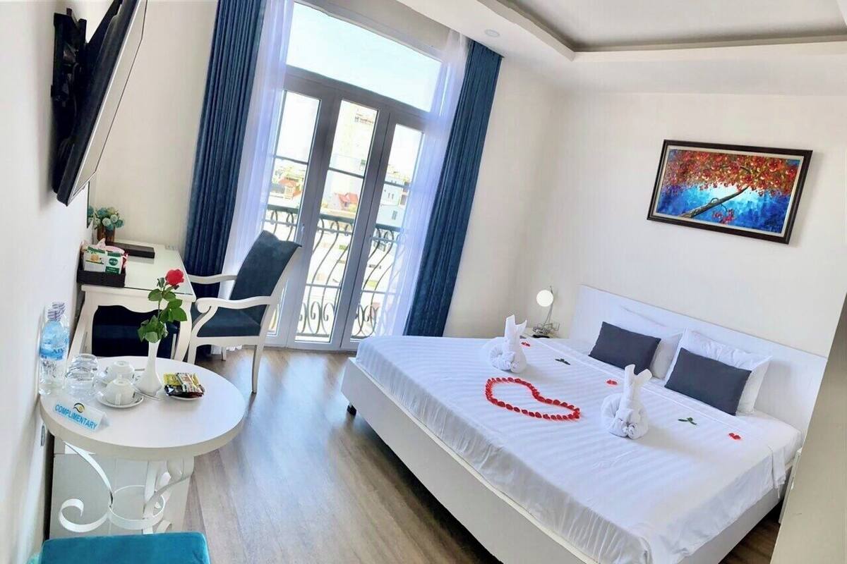A hotel room with a bed, a dining table, chairs, a balcony with blue curtains, and a painting on the wall.