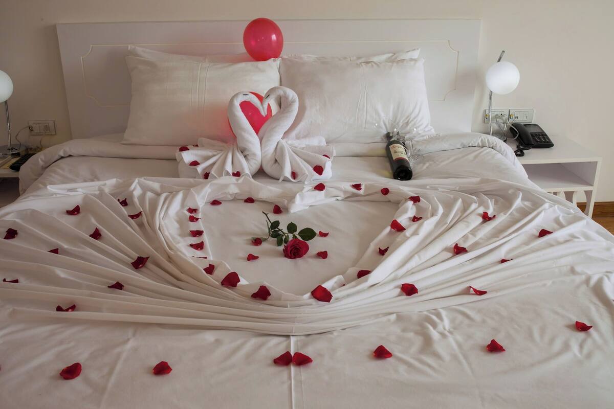 A neatly made bed with a heart-shaped towel arrangement, a bouquet of roses, and a champagne bottle.