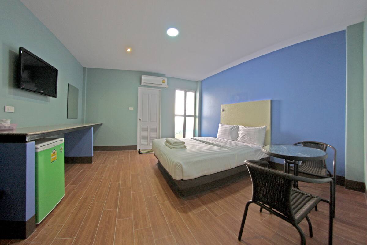 A hotel room with a bed, a desk with a refrigerator, a television, a window, and a small table with chairs.