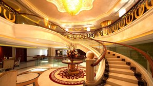 Una majestuosa escalera curva con escalones de mármol y barandilla de cristal, que conduce a un nivel superior con techo decorativo y candelabro.