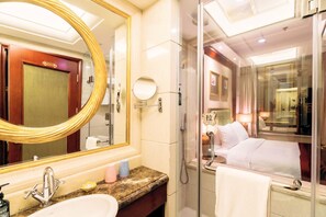 Une salle de bain d’hôtel avec un grand miroir, un lavabo, une douche et un lit visible à travers une paroi en verre.