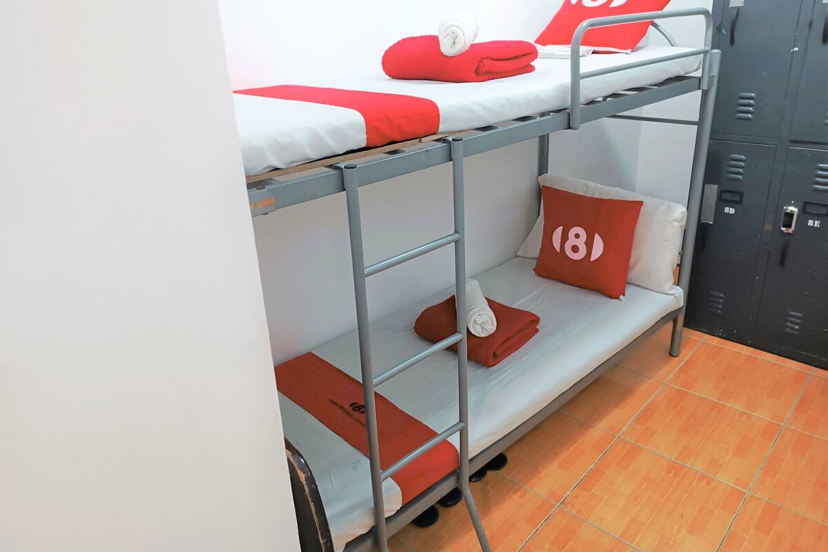 Uma cama beliche com roupa de cama vermelha e branca, um travesseiro vermelho com o número 18, e um conjunto de armários em segundo plano.