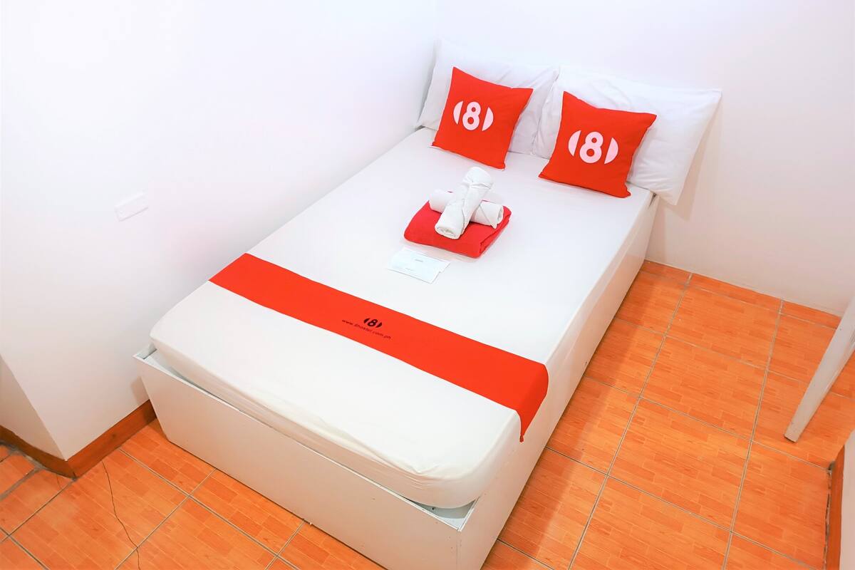 Uma cama bem arrumada com roupa de cama branca e almofadas vermelhas, uma toalha dobrada e um pequeno cartão sobre a cama.