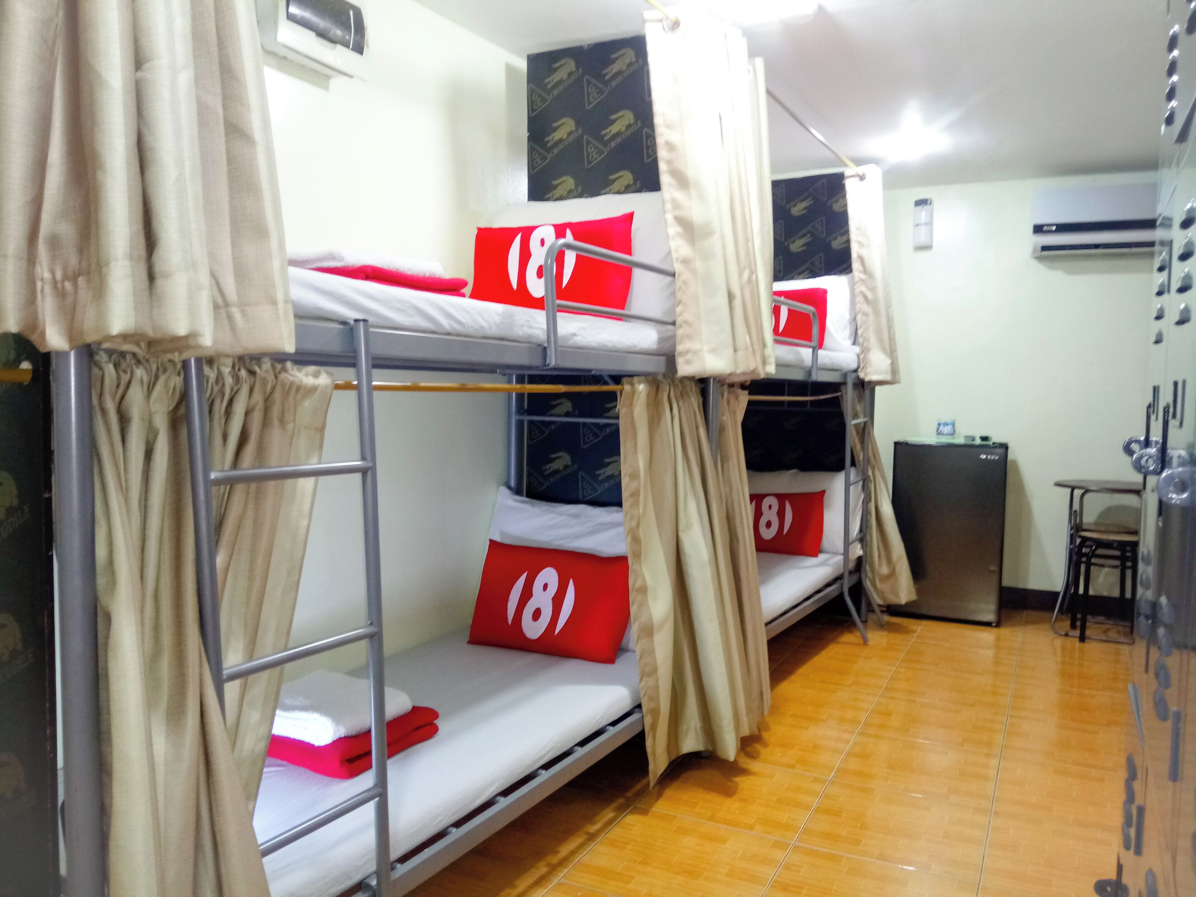 A dormitory room with bunk beds, each with a red pillow and a number tag. There is a small table and a refrigerator in the room.
