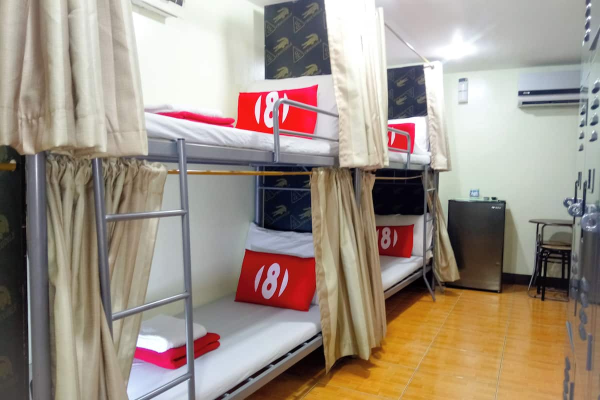 Um quarto de dormitório com beliches, cada um com uma almofada vermelha e uma etiqueta numérica. Há uma pequena mesa e uma geladeira no quarto.