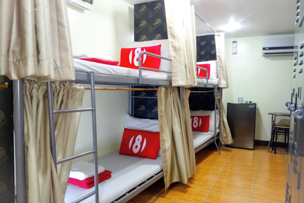 A dormitory room with bunk beds, each with a red pillow and a number tag. There is a small table and a refrigerator in the room.