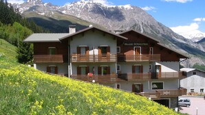 A two-story mountain lodge with balconies, named 'Meublè ROSALPINA', surrounded by a mountainous landscape and a field of yellow flowers. - Meublè Rosalpina (Valdidentro)