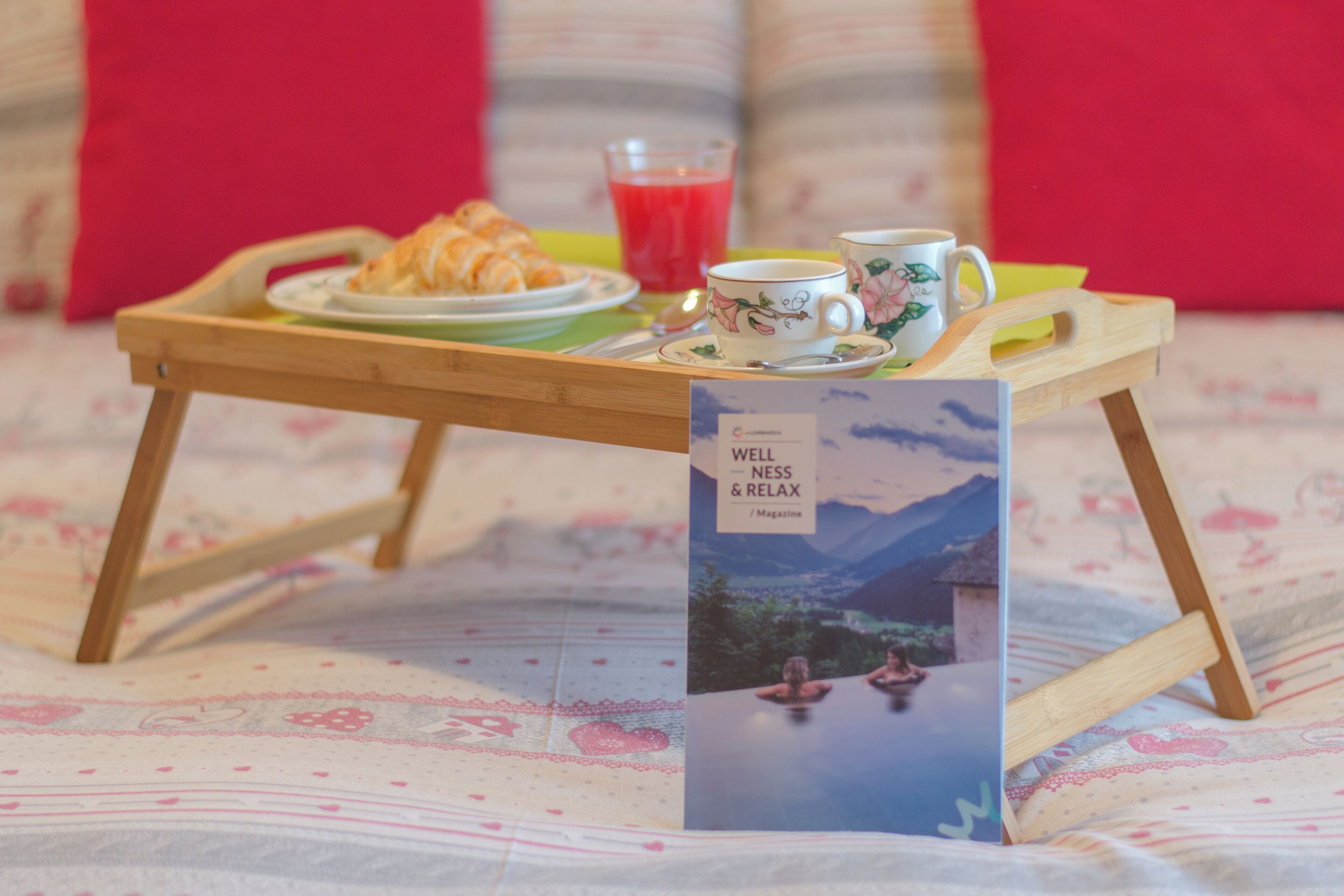 Un plateau en bois avec des croissants, un verre de jus rouge et deux tasses, le tout posé sur un lit recouvert d’une couverture à motifs floraux.