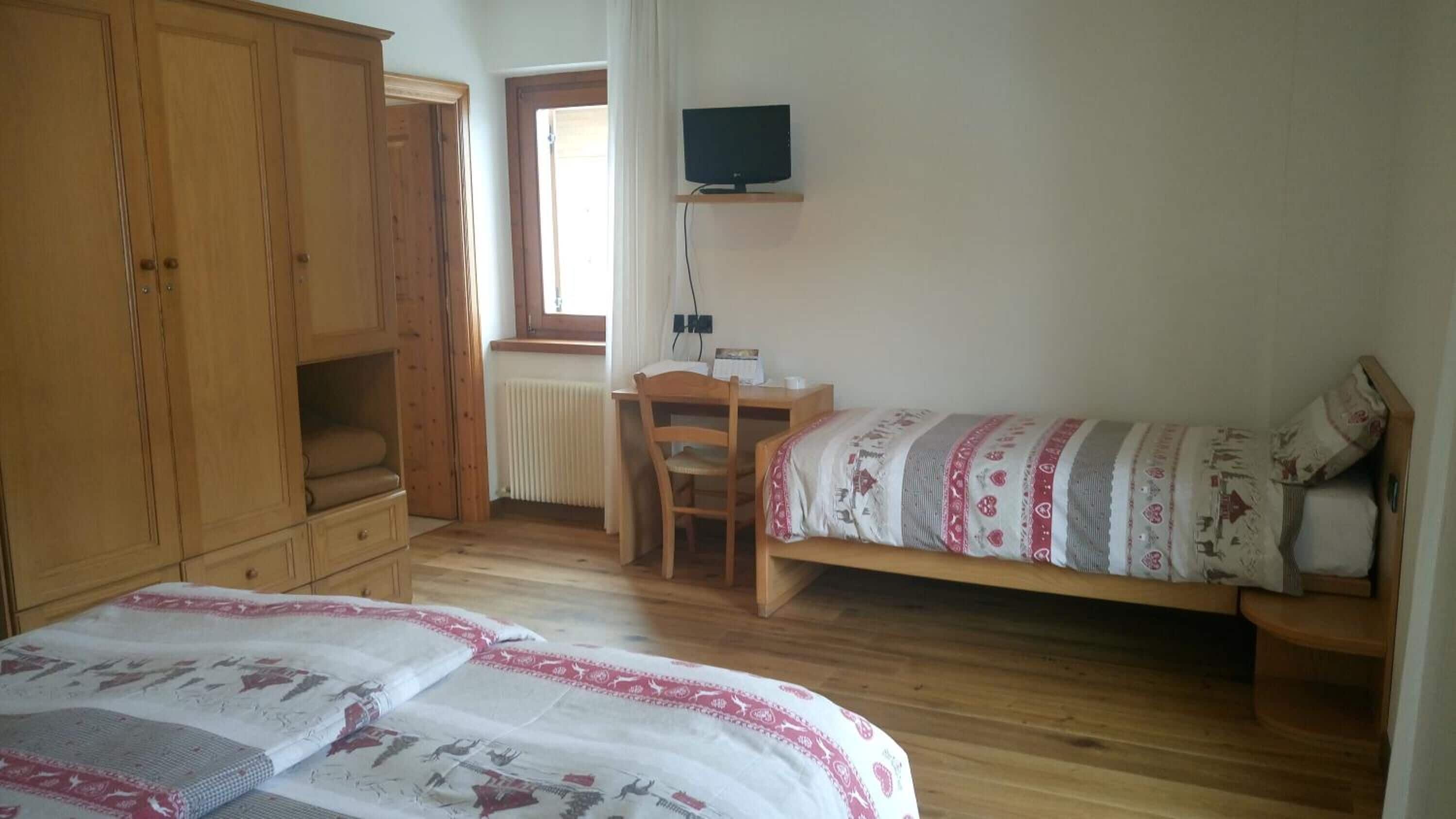 Un dormitorio con dos camas, un armario de madera, un escritorio con una silla, un televisor en una estantería y una ventana con cortinas.