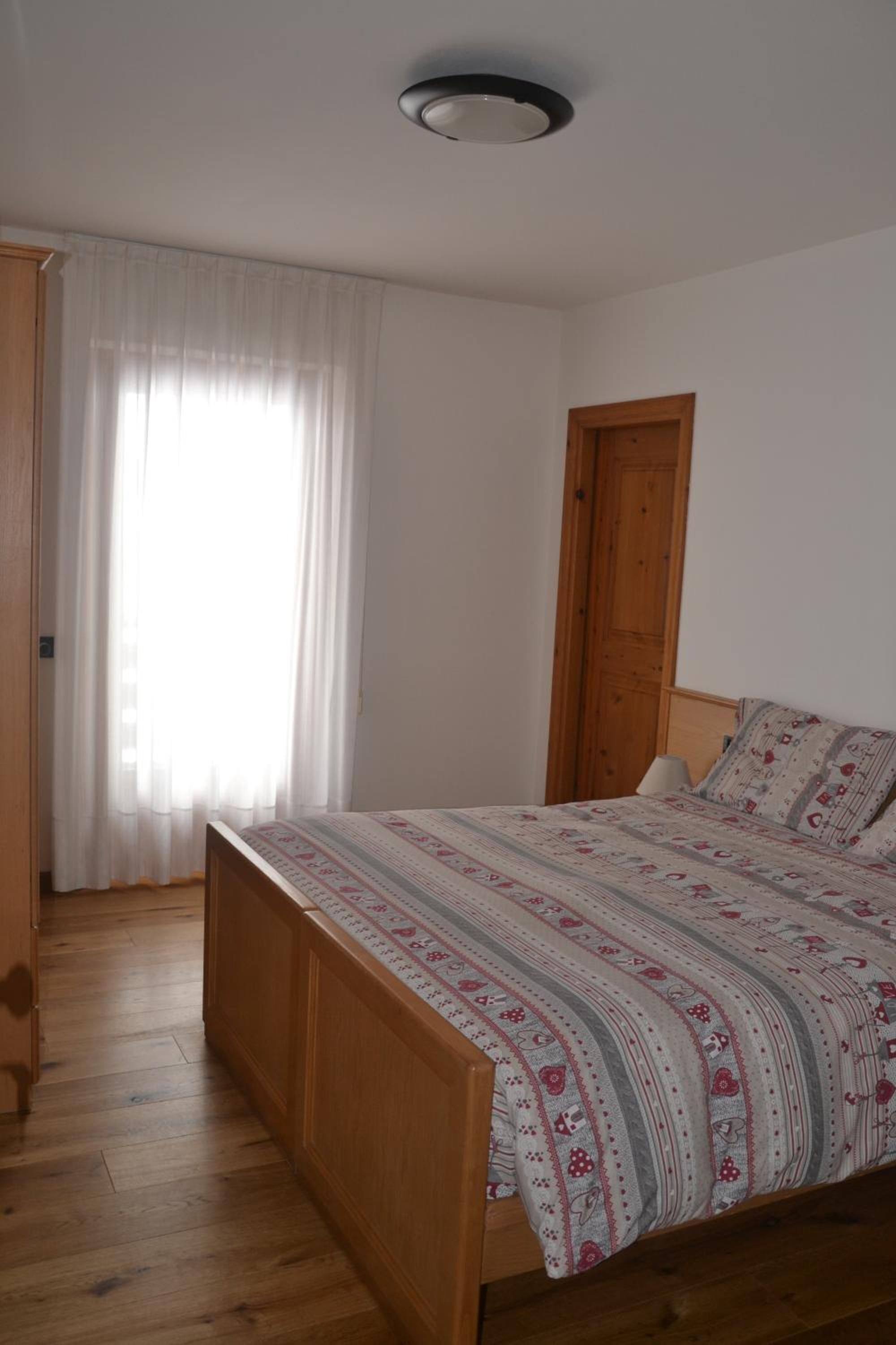 A bedroom with a wooden bed, a wooden wardrobe, a wooden door, and a window with sheer curtains.