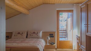 A bedroom with a wooden ceiling, a bed with patterned bedding, a bedside lamp, and a view of the outside through a door. - Meublè Rosalpina (Valdidentro)