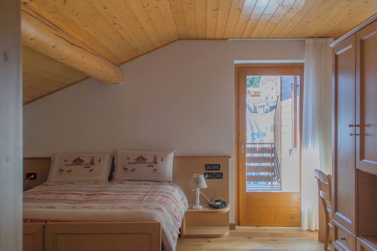 A bedroom with a wooden ceiling, a bed with patterned bedding, a bedside lamp, and a view of the outside through a door.