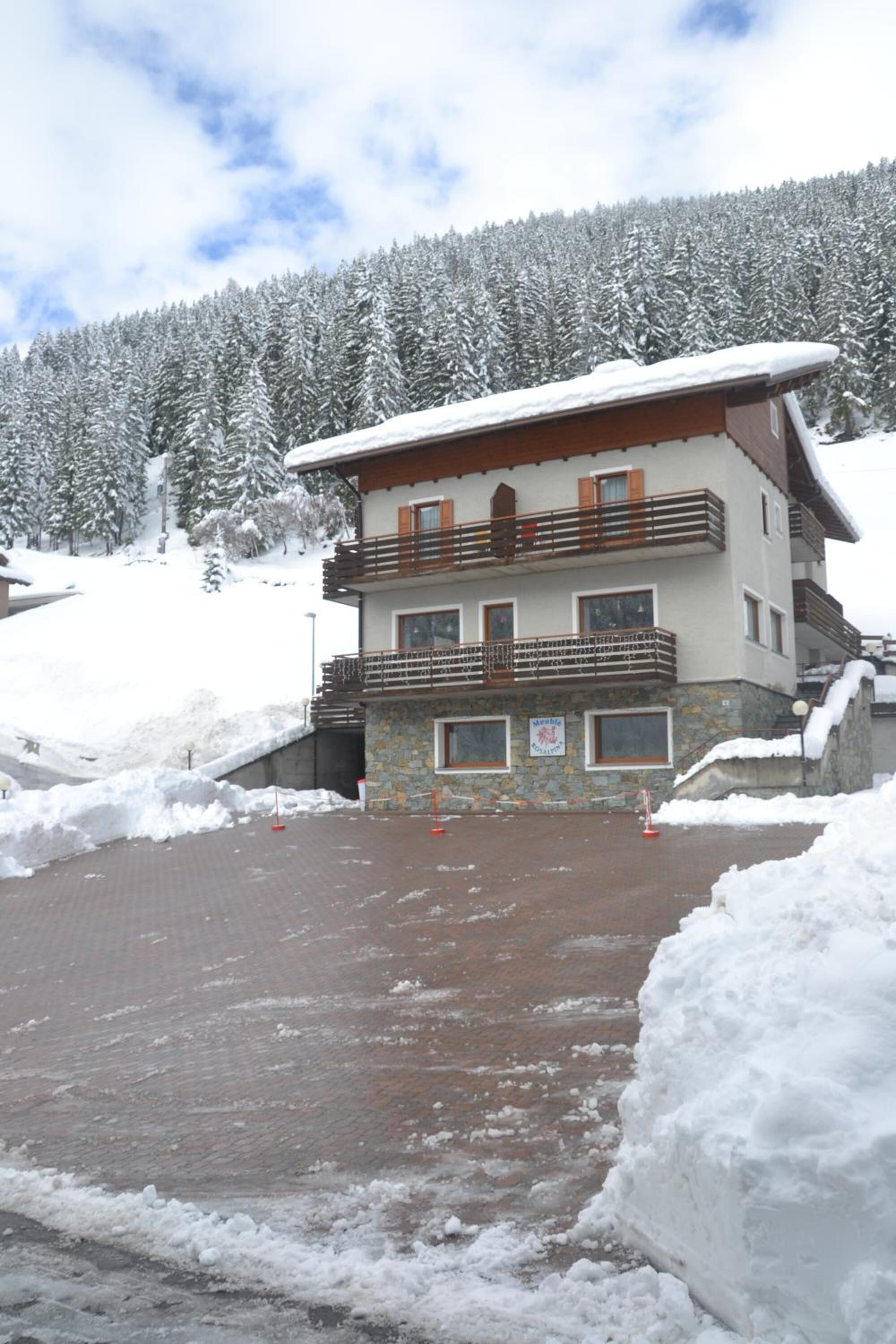 A multi-story building with a snow-covered roof and balconies, surrounded by a snowy landscape and trees.