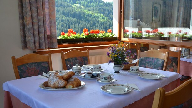 A dining table set with pastries and tea cups, a flower vase, and a window with a view of a forest and flowers. - Meublè Rosalpina (Valdidentro)