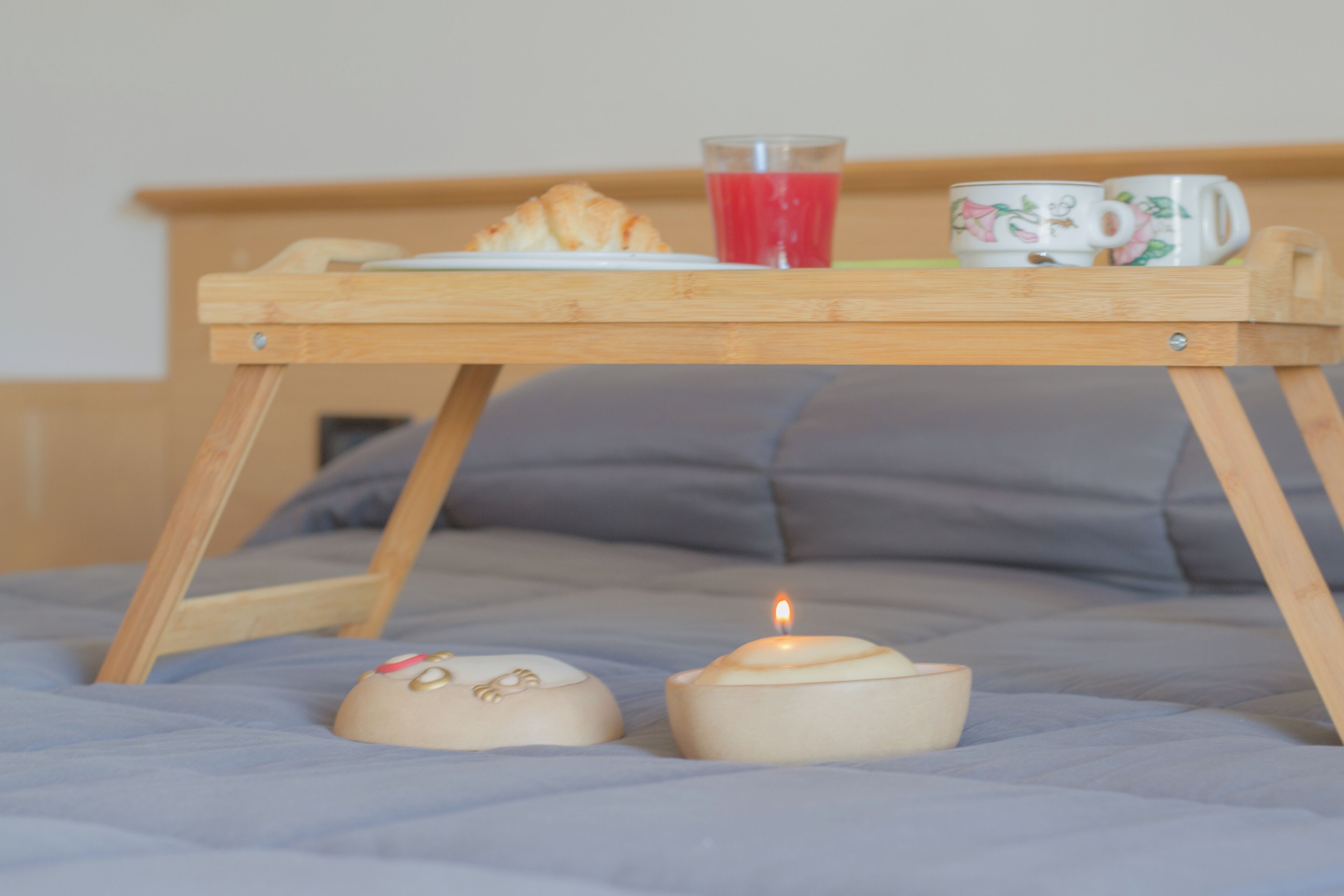 A wooden tray with a croissant, a glass of red juice, and two mugs on a bed.