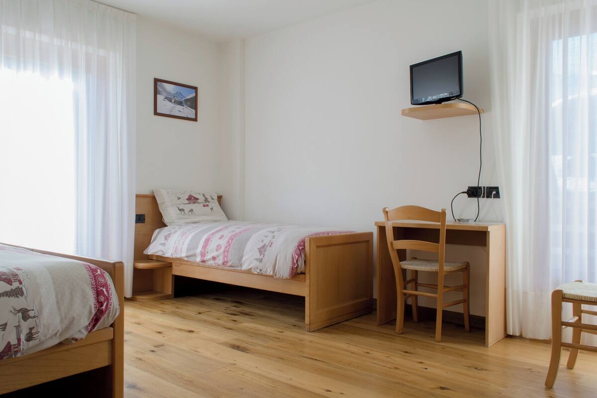 A room with a bed, a desk with a chair, a television on a shelf, and a picture on the wall.