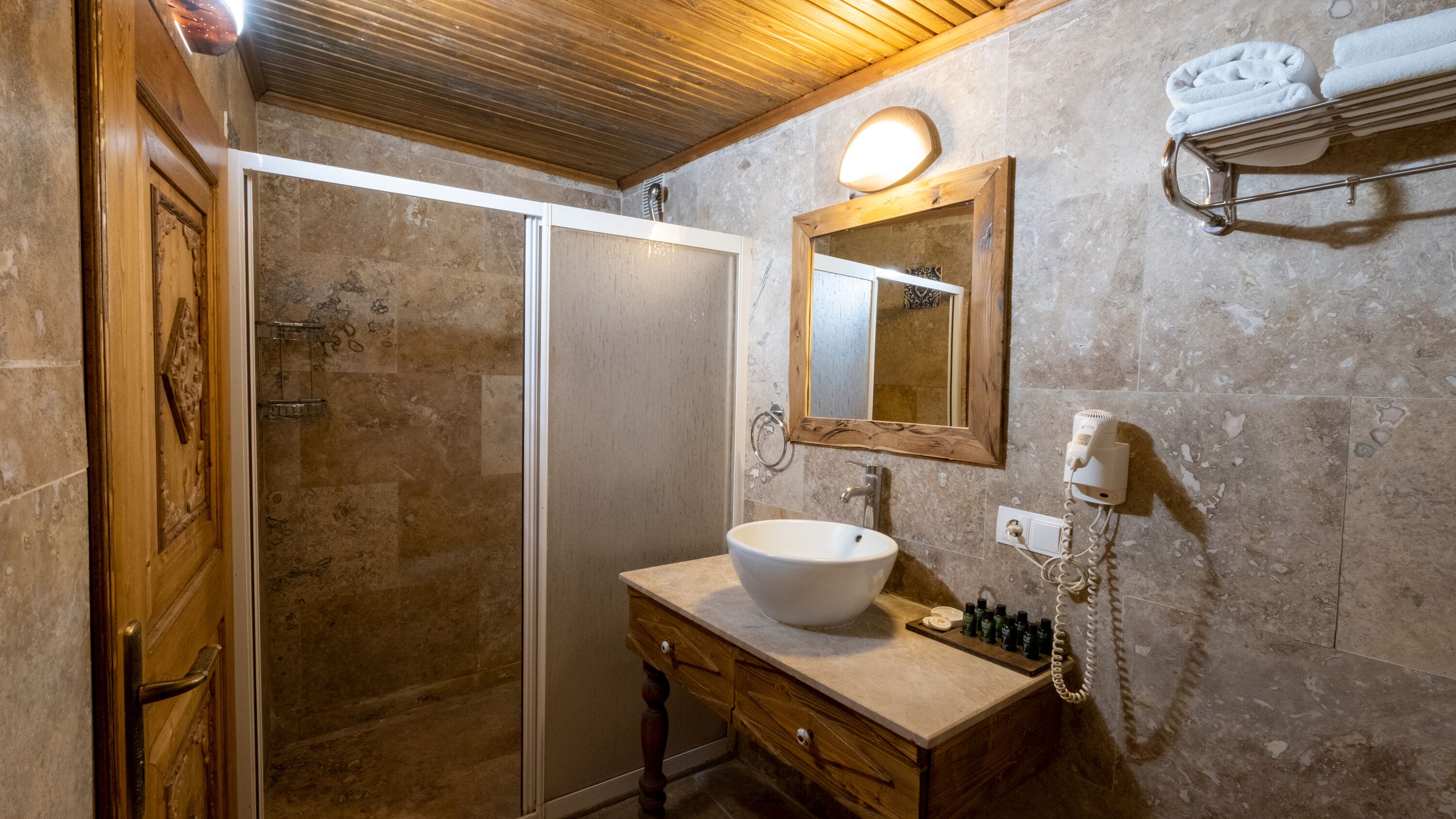 Un baño con puerta de madera, cabina de ducha con mampara de vidrio, lavamanos con espejo y toallero con toallas.