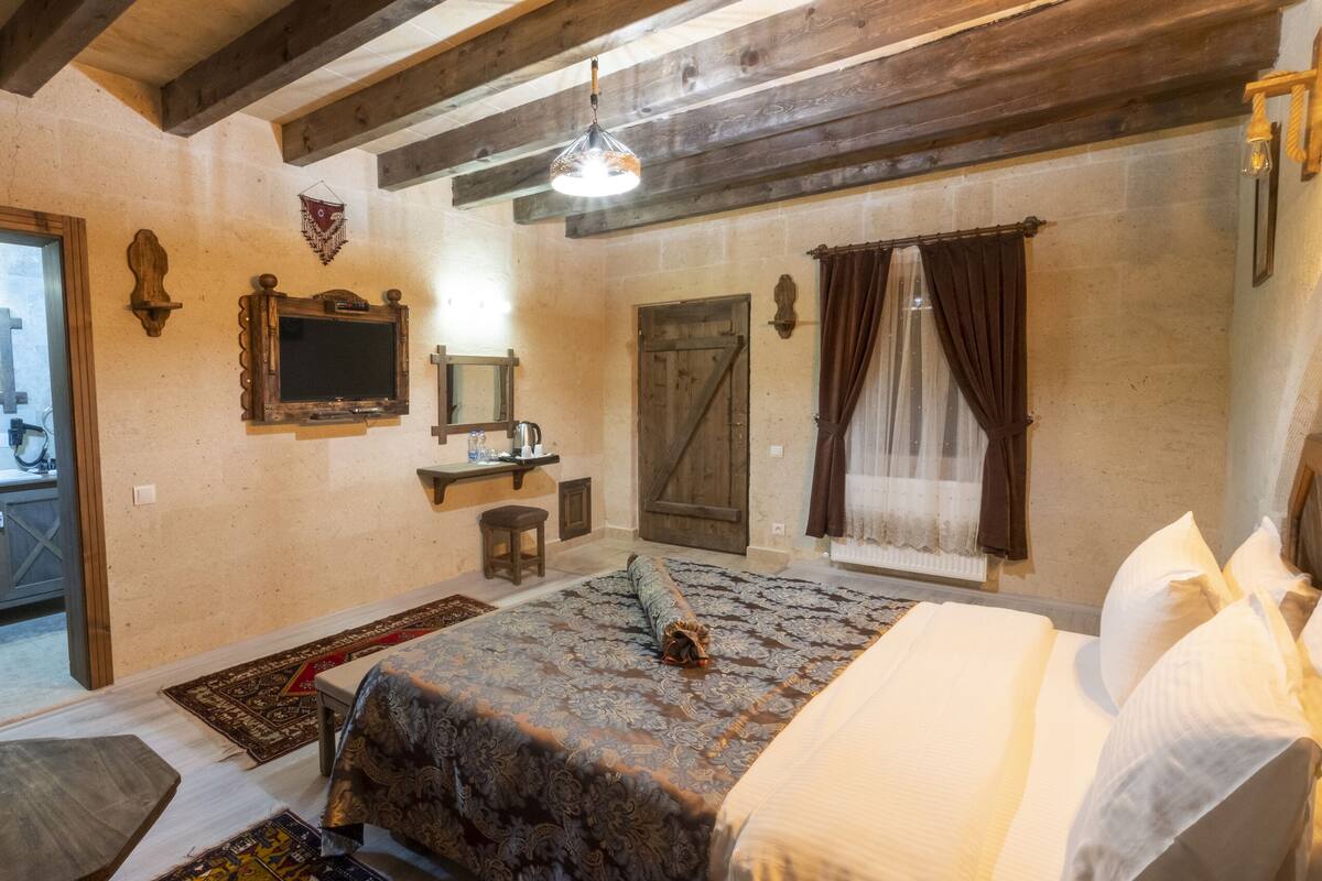 Una habitación rústica con una cama, un banco de madera, una mesita, un espejo, un televisor y una ventana con cortinas.