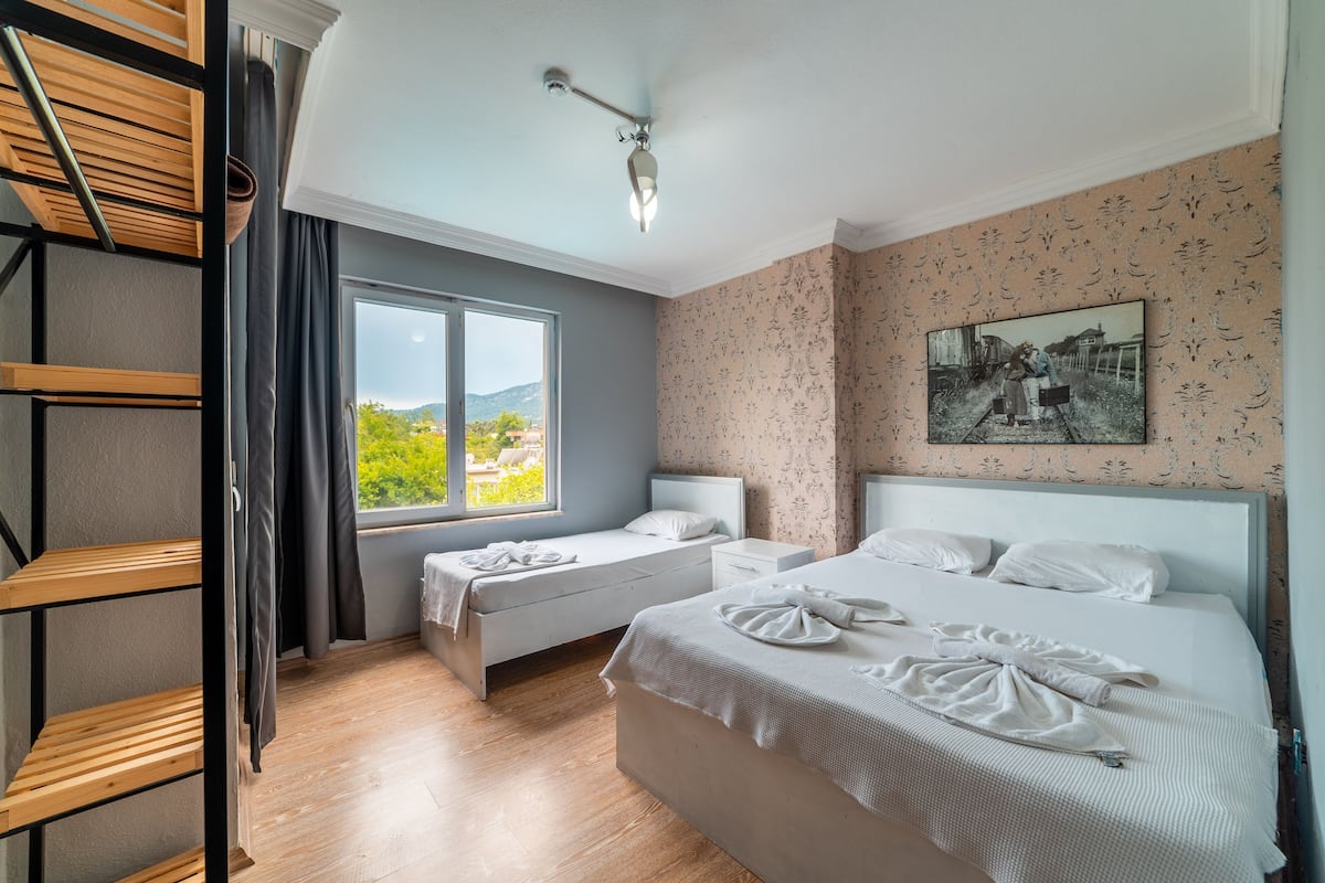 A hotel room with two beds, a window with a view, a wooden shelf, and a painting on the wall.
