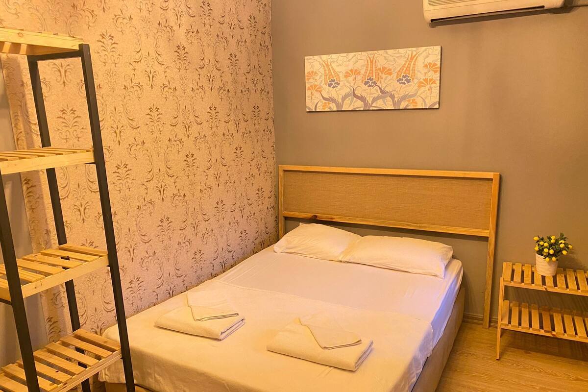 A neatly made bed with a wooden headboard, a wall-mounted air conditioner, and a floral-patterned wallpaper.