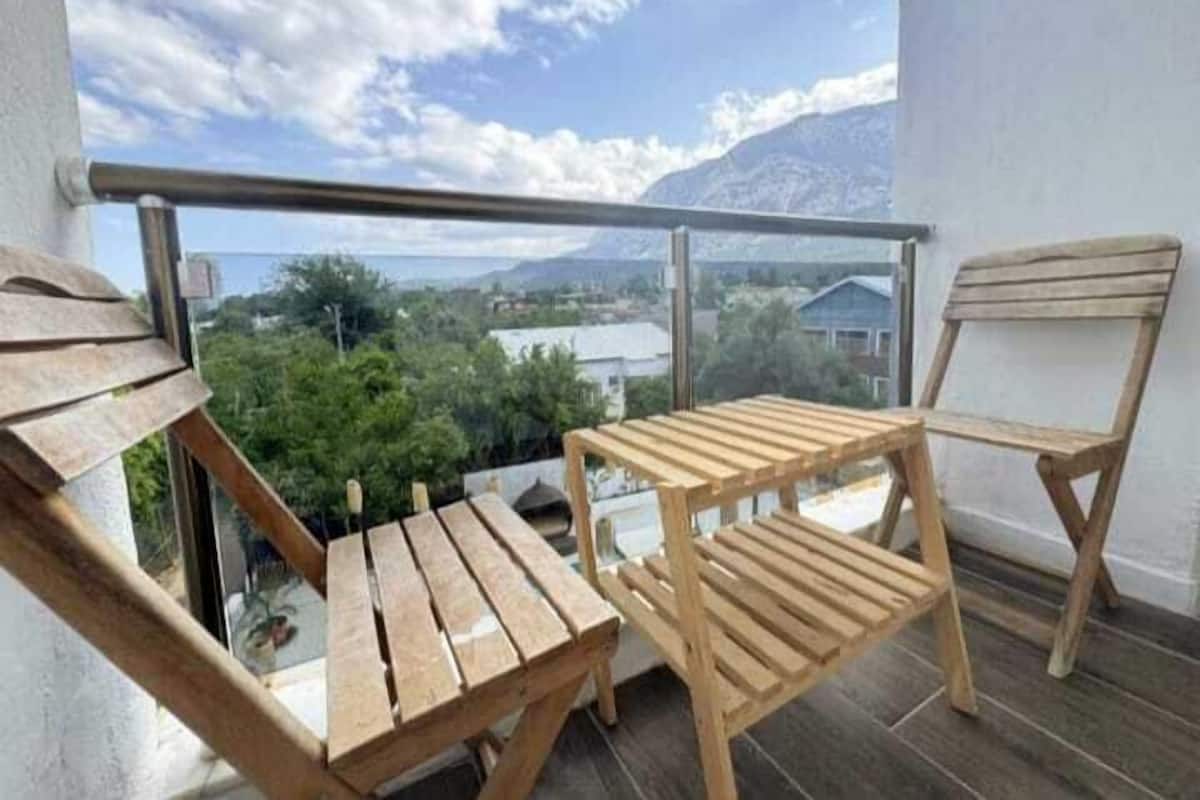 A balcony with wooden furniture, including a table and two chairs, overlooking a scenic view with mountains and houses.