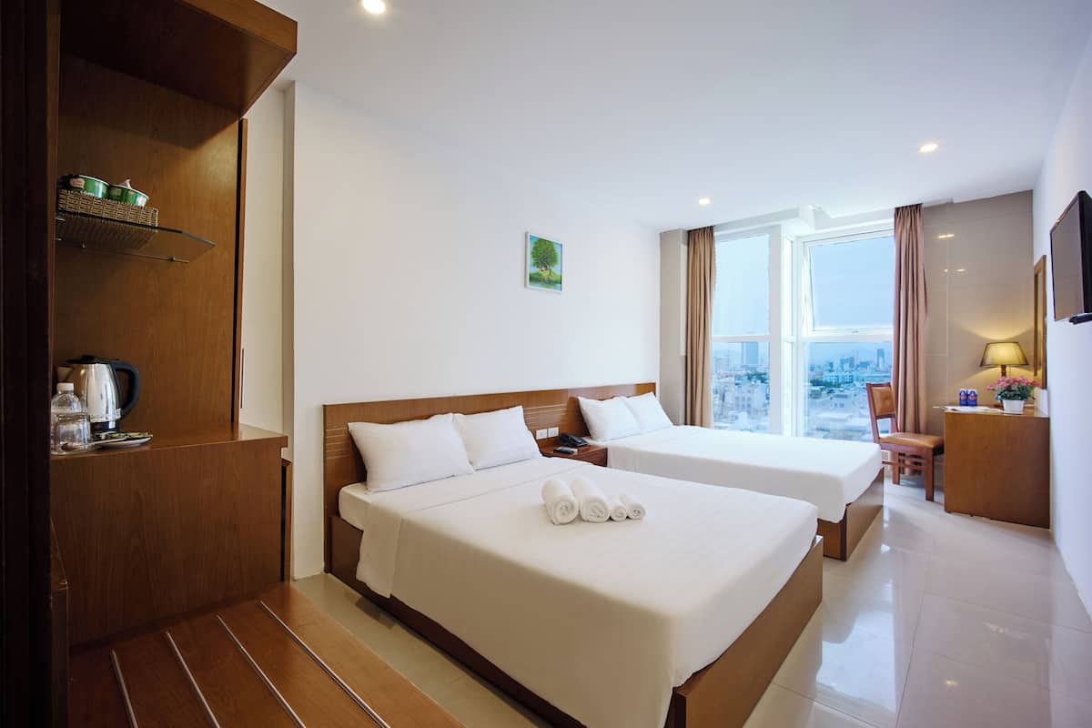 A hotel room with two beds, a wooden cabinet, a window with a city view, and a desk with a lamp.