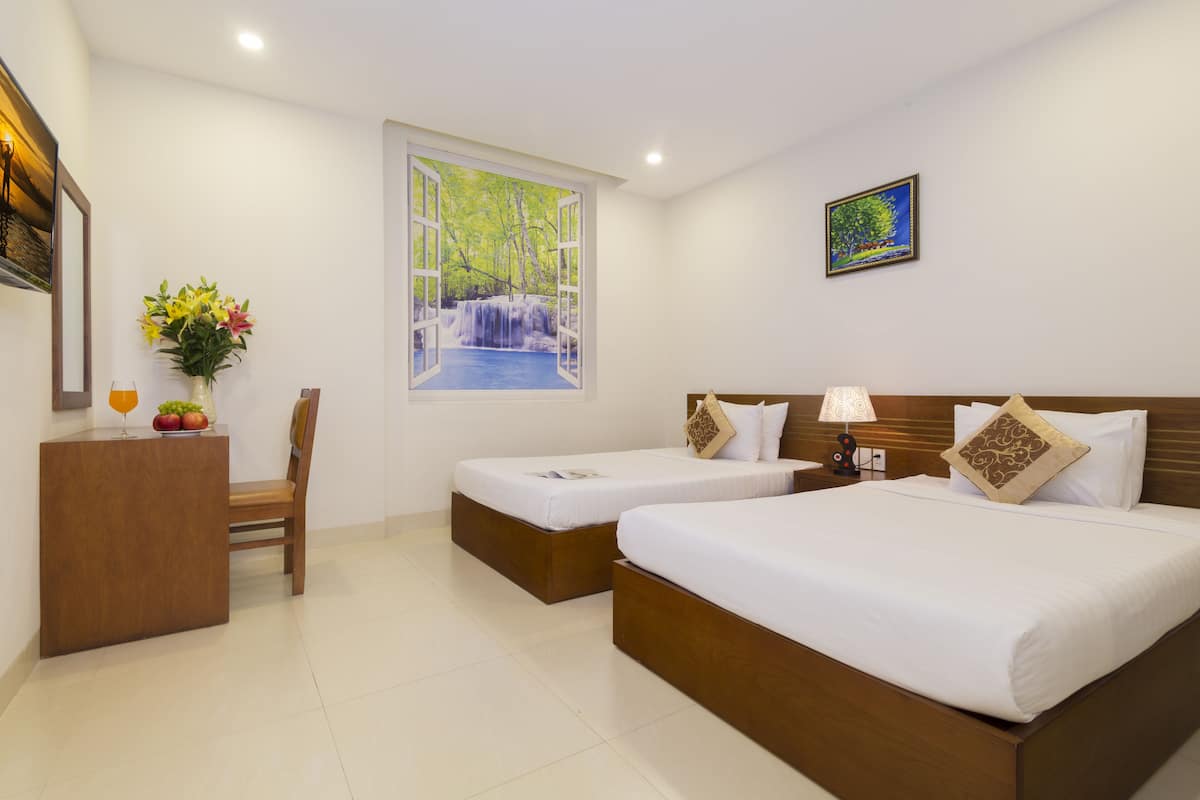 A hotel room with two single beds, a desk with a chair, a vase with flowers, a painting of a waterfall, and a framed artwork on the wall.