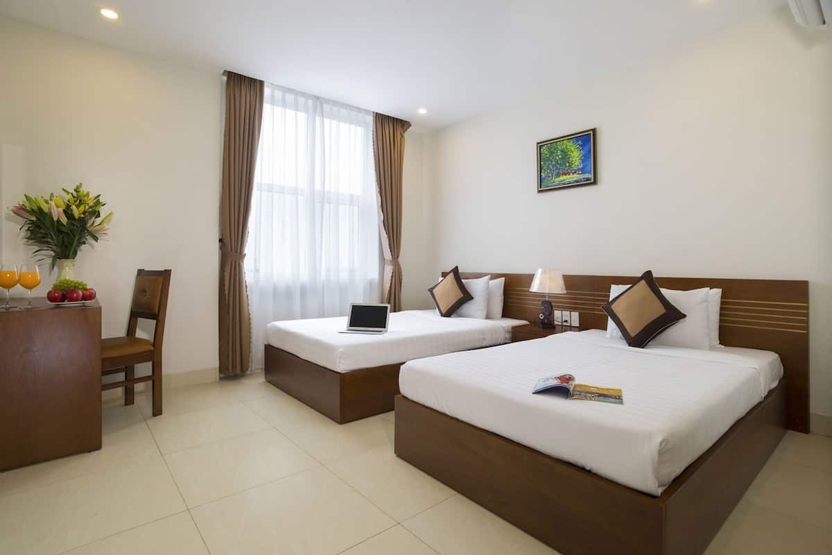 A hotel room with two beds, a desk with a laptop, a chair, a vase with flowers, and a painting on the wall.